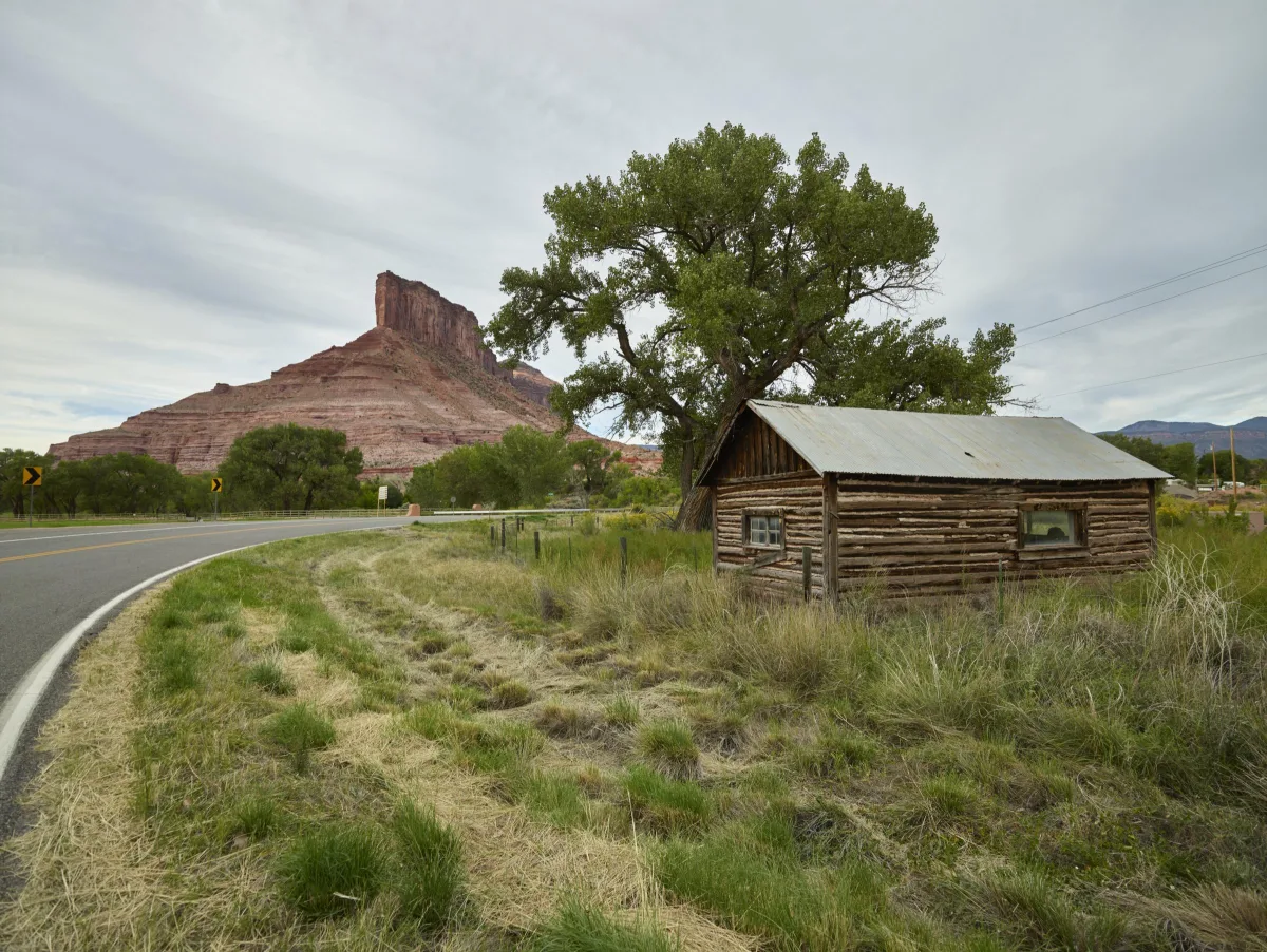 A weathered log cabin in tall grass beside a curving road, below the red rock buttress called The Palisade